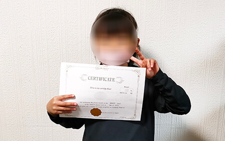 英検Jr.で好成績を収めたことを証明する賞状を手に、ピースサインをする小学1年生の女の子
